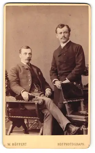 Fotografie W. Höffert, Berlin, Hamburg, Portrait zwei junge Herren in Anzügen auf Stuhl sitzend und an Tisch gelehnt