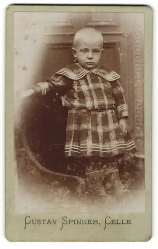Fotografie Gustav Spinner, Celle, Portrait Kind mit geschorenem Kopf in kariertem Kleid