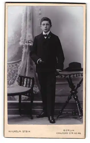 Fotografie Wilhelm Stein, Berlin, Portrait junger Mann mit Fliege u. Handschuhen im Anzug an Tisch gelehnt