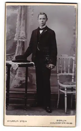 Fotografie Wilhelm Stein, Berlin, Portrait junger Herr mit Seitenscheitel u. Fliege im Anzug an Tisch gelehnt