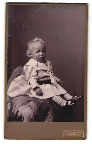 Fotografie J. Kuban, Konstanz, Portrait niedliches Kleinkind im Matrosenkleid auf Fell sitzend