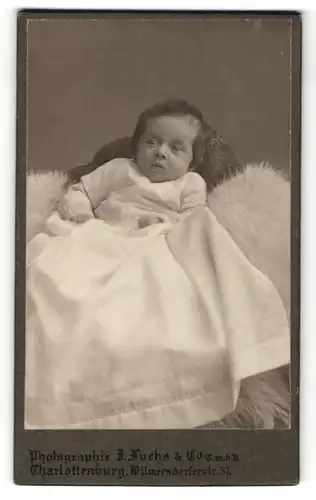 Fotografie J. Fuchs & Co., Charlottenburg, Portrait niedliches Baby im weissen Kleid auf Fell sitzend