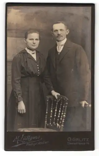 Fotografie M. Lüttgens, Görlitz, Portrait junges Paar in zeitgenössischer Kleidung an Stuhl gelehnt