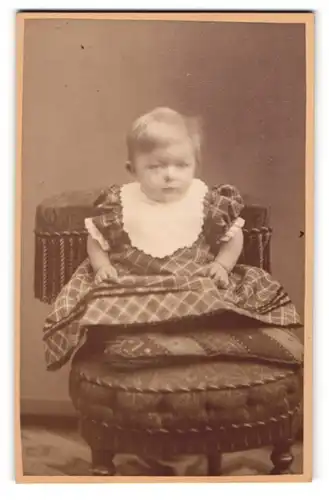 Fotografie Eulenstein, Leipzig, Portrait niedliches Kleinkind mit Latz im karierten Kleid auf Hocker sitzend