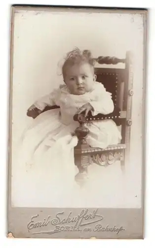 Fotografie Emil Schuffert, Borna, Portrait niedliches Baby im weissen Kleid auf Hochstuhl sitzend