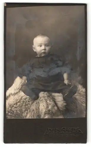 Fotografie Louis Ahrens, Winsen a / d. Luhe, Portrait niedliches Kleinkind im hübschen Kleid auf Fell sitzend
