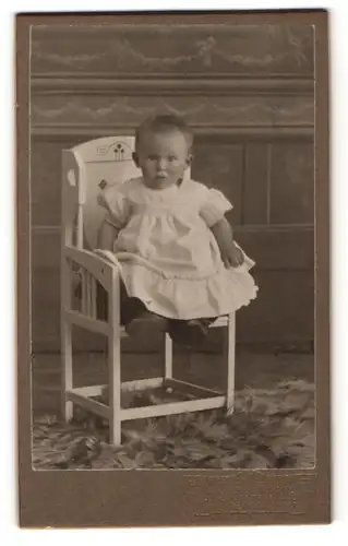 Fotografie Julius Grusche, Neugersdorf i. S., Portrait blondes niedliches Baby im weissen Kleidchen auf einem Kinderstuhl