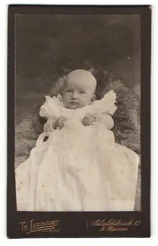 Fotografie Th. Liebert, Bremen-Sebaldsbrück, Portrait niedliches Baby im weissen Kleid auf Fell sitzend