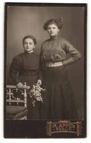 Fotografie M. Appel, Berlin, Portrait zwei junge Damen mit zeitgenöss. Frisuren u. Blumen an Stuhl gelehnt