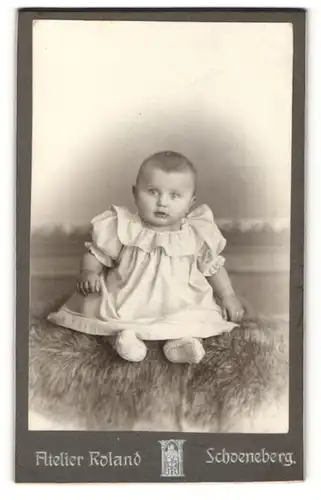 Fotografie Atelier Roland, Berlin-Schoeneberg, Portrait niedliches Baby im hübschen Kleid auf Fell sitzend