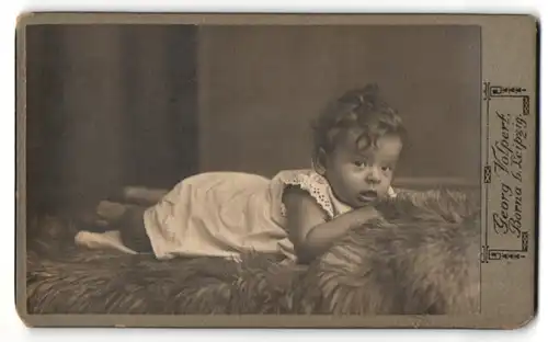 Fotografie Georg Volpert, Borna, Portrait niedliches Kleinkind im weissen Hemd bäuchlings auf Fell liegend