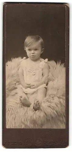 Fotografie J. Fuchs, Schöneberg, Portrait niedliches Kleinkind im weissen Hemd auf Fell sitzend
