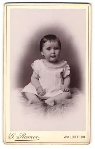 Fotografie G. Ramer, Waldkirch, Portrait niedliches Kleinkind im weissen Kleid auf Fell sitzend