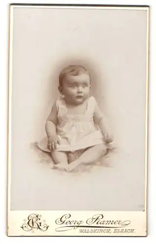 Fotografie Georg Ramer, Waldkirch, Portrait niedliches Baby im weissen Kleid auf Fell sitzend