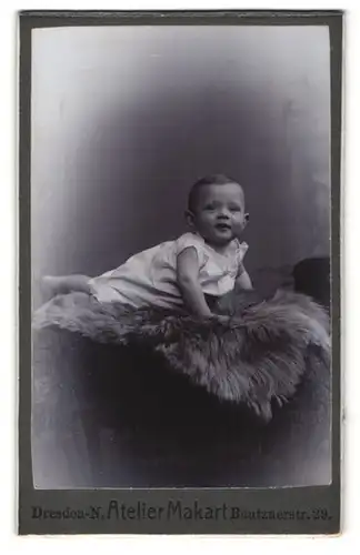 Fotografie Atelier Makart, Dresden-N., Portrait niedliches Baby im weissen Kleid auf Fell liegend