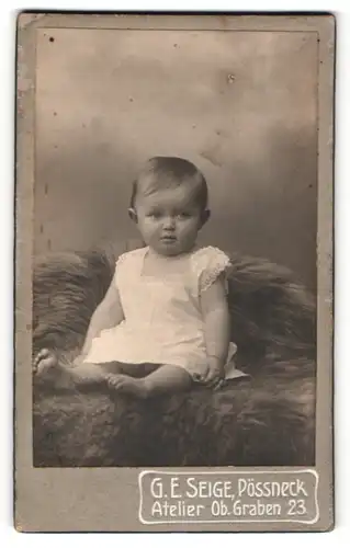 Fotografie G. E. Seige, Pössneck, Portrait niedliches Kleinkind im weissen Kleid auf Fell sitzend