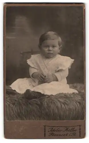 Fotografie Atelier Müller, Pössneck i. Th., Portrait niedliches Kleinkind im weissen Kleid auf Fell sitzend