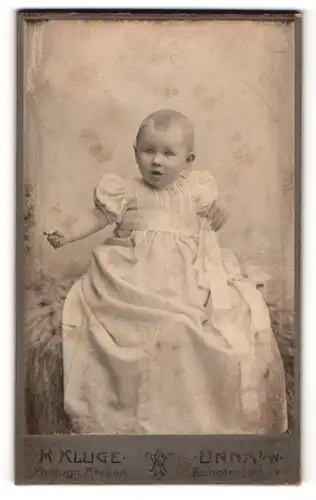 Fotografie H. Kluge, Unna i / W., Portrait niedliches Kleinkind im hübschen Kleid auf Fell sitzend
