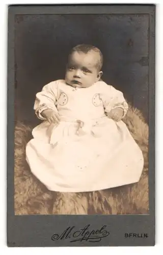 Fotografie M. Appel, Berlin, Portrait niedliches Baby im weissen Kleid auf Fell sitzend
