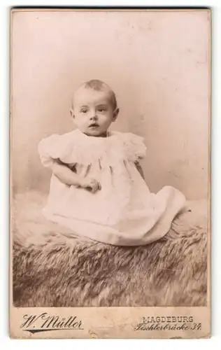 Fotografie W. Müller, Magdeburg, Portrait niedliches Kleinkind im weissen Kleid auf Fell sitzend