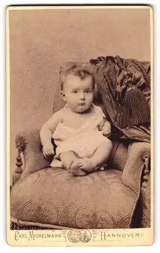 Fotografie Carl Michelmann, Hannover, Portrait niedliches Baby im weissen Hemd auf Sessel sitzend