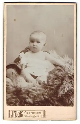 Fotografie J. Fuchs, Berlin-Charlottenburg, Portrait niedliches Baby im weissen Hemd auf Fell sitzend