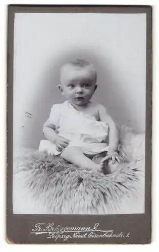 Fotografie Fr. Brüggemann, Leipzig, Portrait niedliches Baby im weissen Hemd auf Fell sitzend