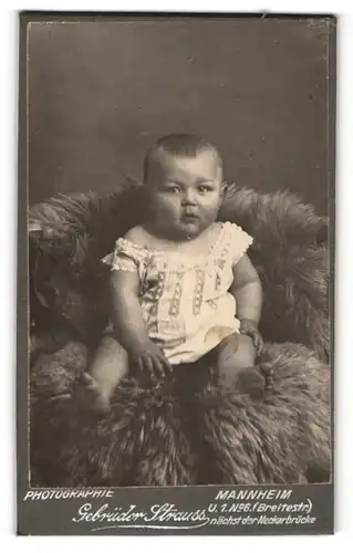 Fotografie Gebrüder Strauss, Mannheim, Portrait niedliches Baby im Hemdchen auf Fell sitzend