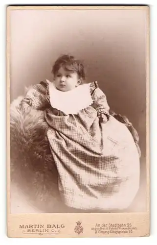 Fotografie Martin Balg, Berlin C., Portrait niedliches Kleinkind im karierten Kleid mit Latz auf Fell sitzend