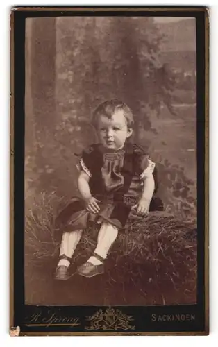 Fotografie R. Spreng, Bad Säckingen, Portrait bezauberndes kleines Mädchen mit Riemenschühchen auf Stroh sitzend