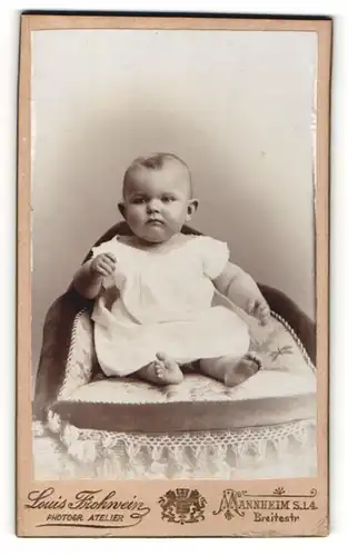 Fotografie Louis Frohnwein, Mannheim, Portrait niedliches Baby im weissen Kleid auf Kissen sitzend
