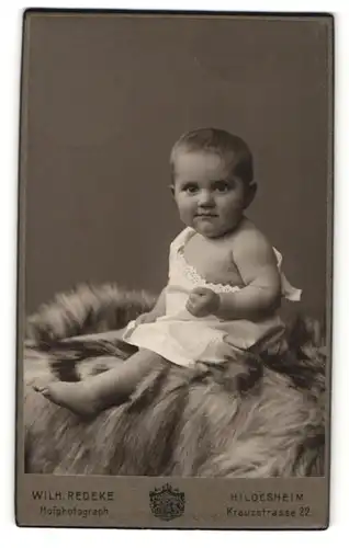 Fotografie Wilh. Redeke, Hildesheim, Portrait niedliches Kleinkind im weissen Hemd auf Fell sitzend