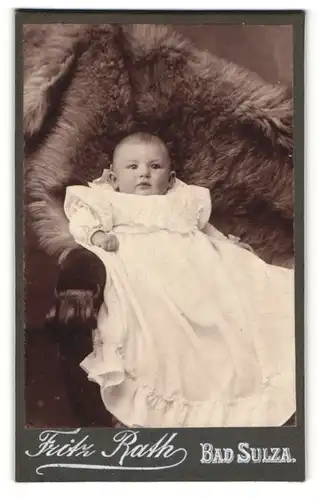 Fotografie Fritz Rath, Bad Sulza, Portrait niedliches Baby im weissen Kleid auf Fell liegend