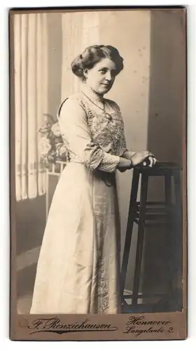 Fotografie F. Renziehausen, Hannover, Portrait lächelnde hübsche Dame im bestickten Kleid am Hocker stehend