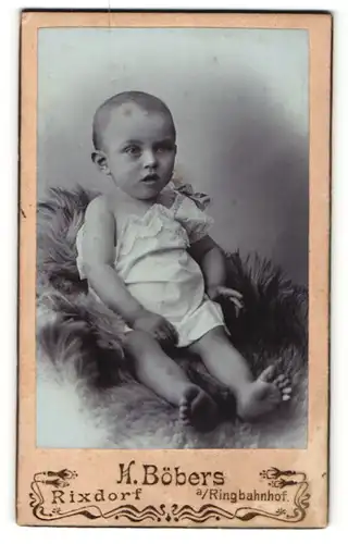Fotografie H. Böbers, Rixdorf, Portrait niedliches Kleinkind im weissen Hemd auf Fell sitzend