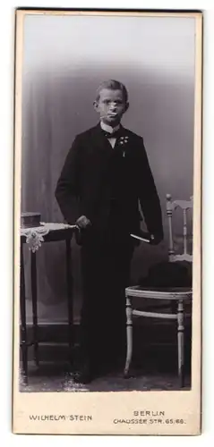 Fotografie Wilhelm Stein, Berlin, Portrait hübscher Knabe im Anzug mit Buch an Tisch gelehnt