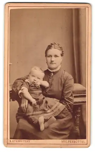 Fotografie A. Sternitzky, Wolfenbüttel, Portrait stolze Mutter in schlichtem Kleid mit Kleinkind im Arm an Lehne Sitzend