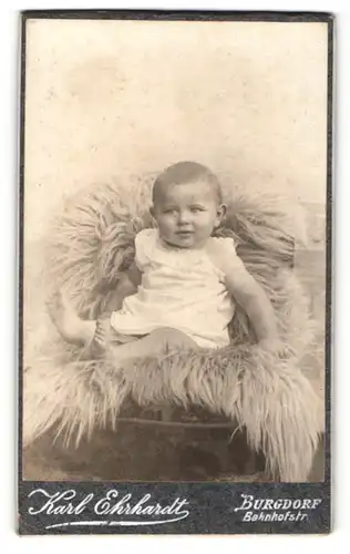 Fotografie Karl Ehrhardt, Burgdorf, Baby im weissen Kleidchen sitzt auf einem Fell