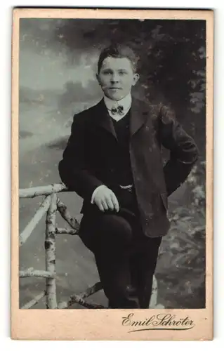 Fotografie Emil Schröter, Jüterbog, Portrait junger Herr mit Fliege im Anzug an Zaun gelehnt