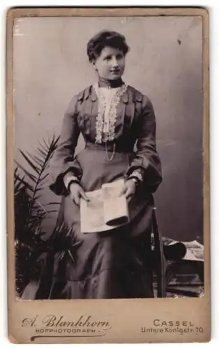 Fotografie A. Blankhorn, Cassel, Portrait junge Dame im eleganten Kleid mit Zeitung an Stuhl gelehnt