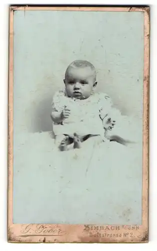 Fotografie G. Göber, Simbach a. Inn, Portrait süsses Baby im weissen Taufkleidchen auf Fell sitzend