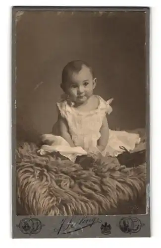 Fotografie J. Seiling, München, Kleinkind im weissen Kleid sitzt auf einem Fellteppich