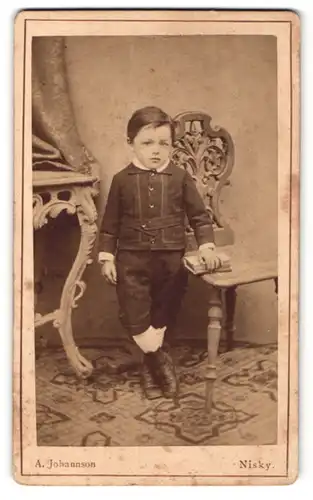 Fotografie A. Johannson, Nisky, Portrait kleiner Junge in hübscher Kleidung mit Buch an Stuhl gelehnt