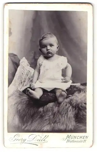 Fotografie Georg Düll, München, Portrait niedliches Kleinkind im weissen Hemd auf Kissen sitzend