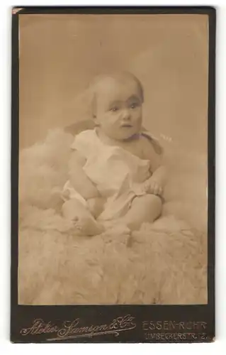 Fotografie Samson & Co, Essen-Ruhr, Portrait niedliches Baby im weissen Hemd auf Fell sitzend