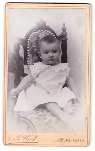 Fotografie M. Wild, Burghausen, Portrait niedliches Kleinkind im weissen Kleid auf Stuhl sitzend