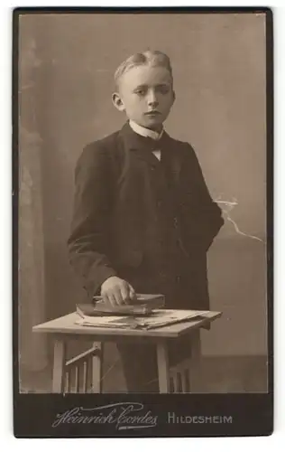 Fotografie Heinrich Cordes, Hildesheim, Portrait frecher blonder Bube im schwarzen Anzug und Buch in der Hand
