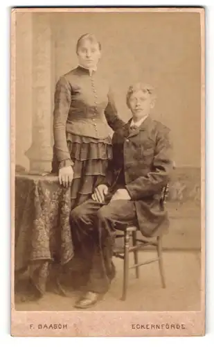 Fotografie F. Baasch, Eckernförde, Portrait bürgerlicher Herr im Anzug am Tisch sitzend u. junge Dame an Tisch gelehnt