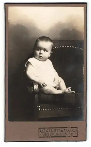 Fotografie Alex. Lauterwasser Jr., Ueberlingen a/B, Portrait Baby im weissen Hemdchen auf einem Sofa sitzend