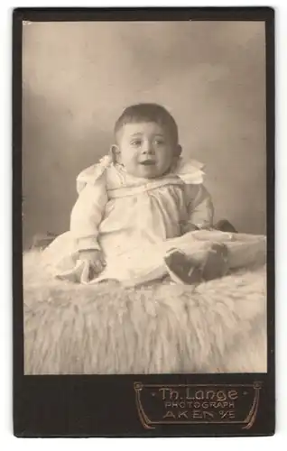 Fotografie Th. Lange, Aken a. E., Portrait lachendes Kleinkind im weissen Kleidchen auf Fell sitzend